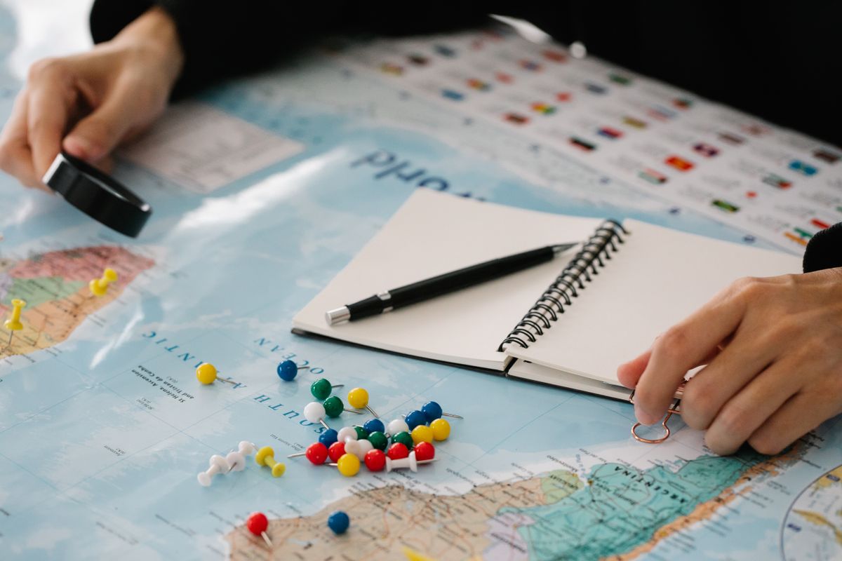 Mãos com lupa, caderno e alfinetes coloridos sobre um mapa-múndi